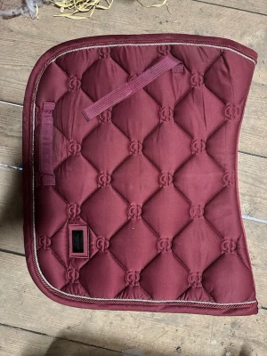Equestrian Stockholm schabrak rött - Säljer ett vinrött schabrak med quiltat mönster och dekorativ kant i beige. Schabraket har praktiska remmar för fastsättning och är tillverkat i ett slitstarkt tyg. Perfekt för dig som vill ha en snygg detalj till hästutrustningen. Det är ett dressyr schabrak i storlek cob, det är använt fåtal gånger och är i mycket bra skick.
