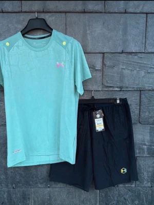 Under Armour träningsset - Snyggt träningsset från Under Armour med en turkos t-shirt i funktionsmaterial och en liten rosa logga på bröstet. Tillhörande svarta shorts med elastisk midja och logga på benet. Perfekt för gym eller löpning.