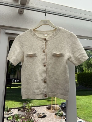 Kortärmad beige kofta med guldknappar - Snygg kortärmad kofta i beige med guldknappar framtill och en bröstficka. Perfekt till både jeans och kjol. Mjuk och lättstickad, passar till många olika stilar. Förliten för mig, därför får denna säljas till en ny ägare☀️