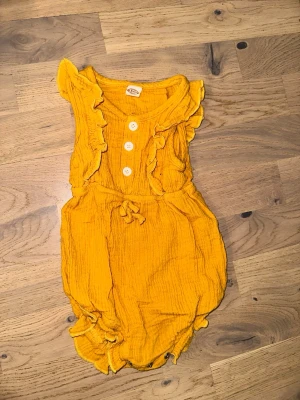 Gul body med volanger och knappar - Supersöt gul body med volangdetaljer längs axlar och ben, samt dekorativa vita knappar framtill. Tillverkad i mjukt, krinklat bomullstyg som känns luftigt och bekvämt. Perfekt för dig som gillar färgglada och lekfulla plagg.Gul body med volanger och knappar