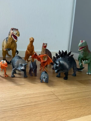 Samling dinosauriefigurer i plast - Säljer en samling med tio olika dinosauriefigurer i plast. Figurerna är i olika färger som grön, orange, svart och grå, och inkluderar både kött- och växtätare samt en flygödla. Perfekt för dig som gillar dinosaurier eller vill ha coola leksaker till samlingen.