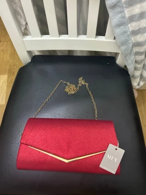 Elegant Röd Kuvertväska med Gulddetaljer – Helt Ny!  - Märke: MUY (Originaletikett kvar).  Färg: Vibrant röd med en sofistikerad lyster.  Design: Klassisk kuvertmodell med en snygg V-formad gulddetalj framtill.  Mångsidighet: Kan bäras som en chic clutch i handen eller som en axelremsväska tack vare den eleganta, avtagbara guldkedjan.  Stängning: Smidig magnetisk tryckknapp som håller dina tillhörigheter säkra.
