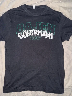 Svart Bajen Södermalm t-shirt - Svart t-shirt med tryck i grönt och vitt där det står 'BAJEN SÖDERMALM 1897' på bröstet. Trycket har en cool graffiti-stil och t-shirten har klassisk passform med rund hals och korta ärmar. Endast använd 2 ggr. Storlek L men sitter som M. Perfekt för dig som vill representera Södermalm och Bajen.