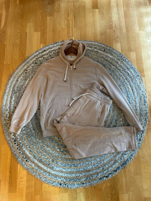 Brunello Cucinelli Tracksuit - Beige loungewear-set med hoodie och matchande byxor ifrån Brunello Cucinelli. Hoodien har dragkedja, fickor och snörning i huvan. Byxorna har resår och dragsko i midjan. Perfekt för chill dagar hemma. Mjukt material för extra komfort.