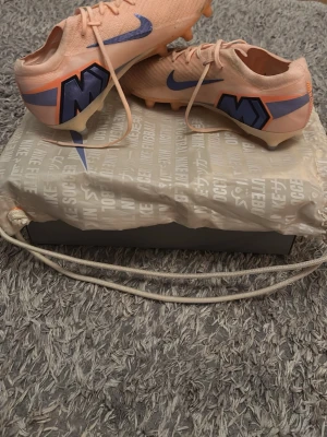 Nike Mercurial rosa fotbollsskor - Säljer ett par Nike Mercurial fotbollsskor i en snygg ljusrosa färg med blå detaljer och stora M-loggor på hälen. Skorna har snörning och är tillverkade i ett lätt syntetmaterial som ger bra bollkontroll och passform. Perfekta för dig som vill sticka ut på planen. Skosnörerna har en knut för de gick av men inget som man märker av, annars är de helt oanvända spelat 2 träningar.