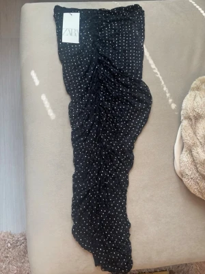 Svart prickig långkjol från Zara - Säljer en svart långkjol från Zara med vita prickar. Kjolen har en åtsittande passform och är lätt rynkad i tyget för en snygg effekt. Perfekt för dig som gillar trendiga och stilrena plagg.