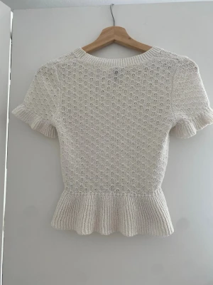 Vit stickad topp med volangkant - Söt vit stickad topp med korta ärmar och volangkant nedtill. Tröjan har ett diskret hålmönster och ribbstickad rund halsringning. Perfekt för dig som gillar en feminin och stilren look.
