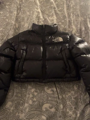 Svart pufferjacka från The North Face - Säljer en svart, glansig pufferjacka från The North Face. Jackan är kortare i modellen, har hög krage och dragkedja framtill. Loggan är broderad i vitt på bröstet. Nypris är 2945.  Priset kan diskuteras!