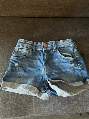 Blå shorts med upprullad kant - Snygga blå shorts med upprullad kant och slitna detaljer framtill. Klassisk ficksmodell med knapp och dragkedja. Perfekta för varma dagar och ger en avslappnad look. Högmidjade och ganska använda säljer för dom blev för små men skulle säga att dom inte ör så slitna.