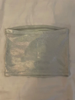 Glittrig silver bandeau tubtopp - Säljer en glittrig silverfärgad bandeau tubtopp som verkligen fångar ljuset. Toppen är axelbandslös och har en tight passform, perfekt för att sticka ut. Materialet känns mjukt och stretchigt, vilket gör den bekväm att bära hela dagen.