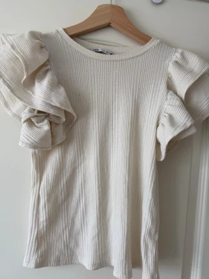 Ribbad volangtop från Zara, vit - Supersnygg ribbad topp från Zara i vit färg med volangdetaljer på ärmarna. Toppen har rund halsringning och är tillverkad i ett mjukt, stretchigt material som sitter bekvämt. Perfekt för dig som vill ha en trendig och unik look.
