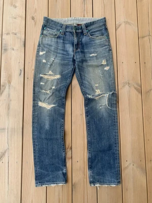 Levi's 506 Distressed - Blå jeans från Levi's med raka ben, slitna detaljer och hål på framsidan för en riktigt edgy look. Klassisk femficksmodell, läderpatch bak i midjan och tillverkade i bomull. Perfekta för dig som gillar en avslappnad och trendig stil.