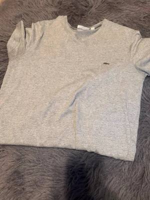 Grå t-shirt från Lacoste - Snygg grå t-shirt från Lacoste med klassisk passform och diskret krokodillogga på bröstet. T-shirten är kortärmad och tillverkad i mjuk bomull, perfekt för en clean och stilren look.