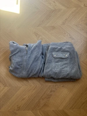 Grått velourset från Juicy Couture - Mysigt grått loungewear-set i velour från Juicy Couture. Setet består av en zip-hoodie med fickor och matchande byxor med resår i midjan och fickor med lock. Perfekt för chill dagar hemma eller när du vill vara bekvämt stylish.