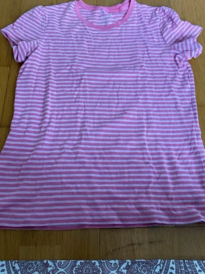 Rosa och vit randig t-shirt - Säljer en söt rosa och vit randig t-shirt med rund hals och korta ärmar. Perfekt för dig som gillar en enkel men ändå färgglad stil. Tillverkad i mjuk bomull som känns skön mot huden. Passar till både jeans och shorts.