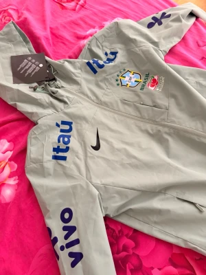 Nike Brasil vindjacka med huva - Ljusgrå vindjacka från Nike med huva och dragkedja. Jackan har Brasilien fotbollslandslagets logga på bröstet, samt blåa tryck med sponsorloggor på ärmar och bröst. Materialet är tunt och vindtätt, perfekt för sportiga dagar. Snygg och sportig design med svarta och blå detaljer.