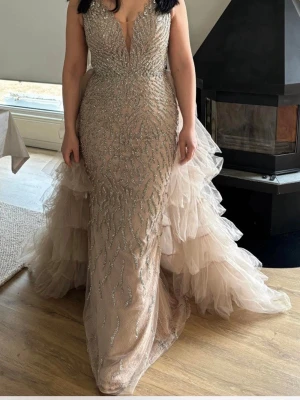 Champagnefärgad långklänning med glitter - Elegant långklänning i champagnefärg med djup v-ringning och glittriga detaljer framtill. Klänningen har volangkjol i flera lager tyll och öppen rygg. Perfekt för bal eller festliga tillfällen. Köpt från sasha brud och fest och använd i endast ett par timmar. Köpt för 4000 och är i väldigt bra skick. Man kan även ta bort veilen där bak och ha den utan.