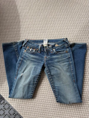 Blå bootcut jeans från True Religion - Säljer ett par blå bootcut jeans från True Religion med slitningar och kontrastsömmar. Jeansen har fem fickor, dekorativa knappar och klassiska ficklock. Skriv för fler frågor och prisförslag.
