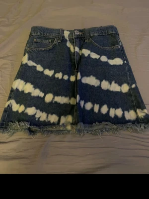 Blå jeanskjol med batikmönster - Cool blå jeanskjol med vitt batikmönster och rå fransig kant nedtill. Kjolen har klassiska fem fickor och knappgylf. Perfekt för dig som vill sticka ut med en unik denimstil.
