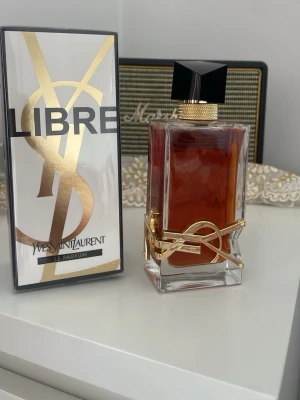 Yves Saint Laurent Libre Le Parfum - Yves Saint Laurent Libre Le Parfum med elegant rektangulär glasflaska, dekorerad med en guldig YSL-logga och svart geometriskt lock. Doften har en djup bärnstensfärg och förpackningen är stilren med guldiga detaljer. Perfekt för dig som gillar lyxiga och moderna parfymer.