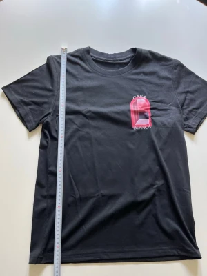 Svart Casa Blanca t-shirt - Snygg svart t-shirt från Casa Blanca med korta ärmar och rund hals. Tryck i rosa och vitt på bröstet med texten 'Casa Blanca' och en grafisk design. Tillverkad i mjuk bomull för en bekväm känsla. Perfekt till jeans eller shorts. Storlek M passar S, nyskick.
