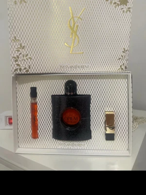 Yves Saint Laurent Black Opium presentset - Lyxigt presentset från Yves Saint Laurent med Black Opium parfym i svart glittrig flaska, matchande miniflaska och ett elegant guldigt läppstift. Förpackningen är vit med gulddetaljer och YSL-logga. Perfekt för dig som gillar exklusiva dofter och snygg design.