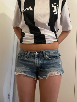 Blå jeansshorts med slitningar - Säljer ett par klassiska blå jeansshorts med slitna detaljer och fransig kant. Shortsen har fem fickor, bälteshällor och knappgylf. Perfekta för varma dagar och ger en avslappnad look.