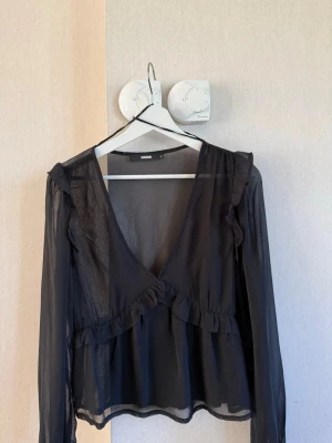 Svart transparent blus med volanger - Snygg svart blus från bik bok i transparent material. Djup V-ringning, volangdetaljer längs bröstet och axlarna samt långa ärmar. Perfekt att styla med ett linne under för en trendig look. Använd fåtal gånger . 