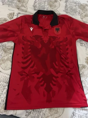 Albaniens fotbollströja röd Macron - Snygg röd fotbollströja från Macron med Albaniens landslagsemblem på bröstet och ett stort örnmönster över hela framsidan. Tröjan har svart krage och svarta detaljer längs sidorna. Perfekt för dig som vill stötta Albanien på match eller ha nått vackert och fint på dig 