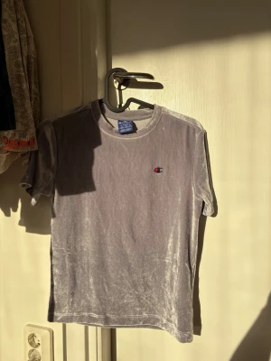 Grå sammets t-shirt från Champion - Säljer en grå sammets t-shirt från Champion med klassisk logga broderad på bröstet. T-shirten har korta ärmar och rund halsringning. Perfekt för dig som vill ha en unik och mjuk t-shirt med lite lyxigare känsla.