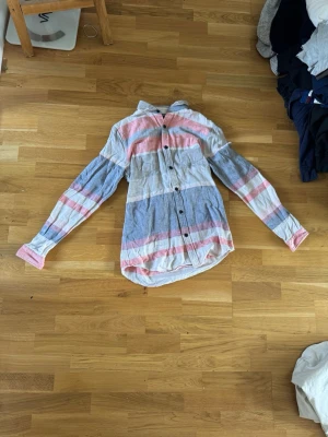 Randig skjorta från Scotch & Soda  - Säljer en långärmad skjorta från Scotch & Soda i storlek M. Skjortan har breda ränder i blått, rosa och vitt, knappar framtill och klassisk krage. Tillverkad i mjuk bomull, perfekt för en avslappnad men snygg look.