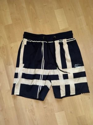 Marinblå och vita mönstrade shorts - Säljer ett par marinblå shorts med breda vita ränder i ett rutigt mönster. Shortsen har resårmidja med snörning och sidofickor. Perfekta för sommaren och har en sportig vibe.