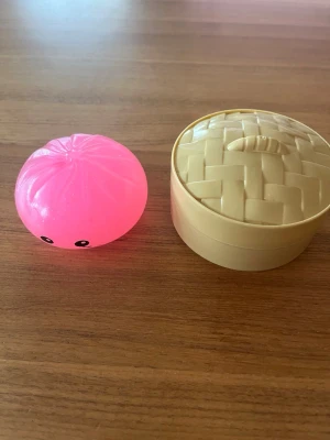 Squishy dumpling rosa glittrig - Rolig squishy dumpling som en glittrig rosa med söta ögon, tillsammans med en beige plastkorg som ser ut som en ångkorg. Perfekt som stressboll eller dekoration på skrivbordet. Leksaken är gjord av mjukt gummi och plast. Väldigt ny. 