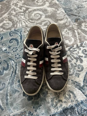 Svarta Moncler Monaco sneakers i mocka storlek 40 - Snygga Moncler Monaco sneakers i svart mocka med vita snören och klassiska Moncler-ränder i rött, vitt och blått på sidan. Låg modell med Moncler-logga på plösen och bekväm innersula. Perfekta för dig som vill ha en stilren och exklusiv look. Skorna är i storlek 40