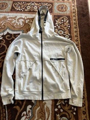 Vit hoodie från Sail Racing - Snygg vit hoodie från Sail Racing med dragkedja framtill och svart logotypdetalj på bröstet. Hoodien har två snedställda fickor, muddar vid ärmslut och en justerbar huva med svarta detaljer. Tillverkad i mjukt bomullsmaterial som känns skönt mot huden.