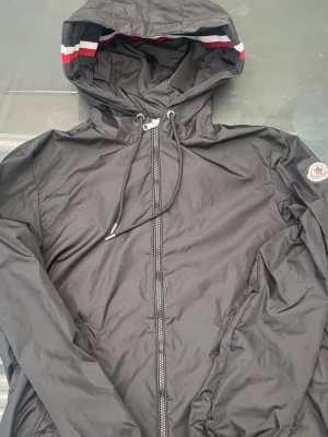 Svart vindjacka från Moncler - Snygg svart vindjacka från Moncler med huva och dragkedja framtill. Jackan har vita och röda detaljer på huvan samt Moncler-logga på ärmen. Tillverkad i lätt och tåligt material, perfekt för blåsiga dagar. Justerbar huva med snörning och två sidofickor.