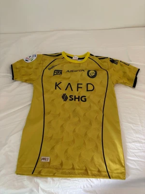 Gul Al Nassr fotbollströja Ronaldo 7 - Snygg gul matchtröja från Al Nassr med svarta detaljer och tryck. Framsidan har klubbmärke, sponsorloggor och texten KAFD SHG. Baksidan har namnet Ronaldo och nummer 7 i svart. Tillverkad i lätt och ventilerande material, perfekt för fotboll eller träning.