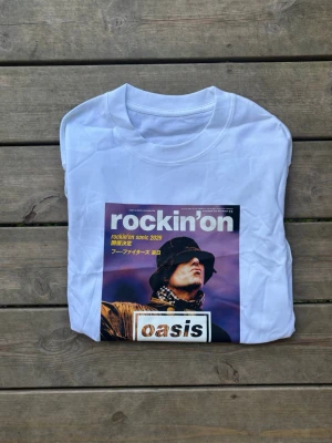 Vit Oasis t-shirt med tryck - Snygg vit t-shirt från rockin'on med stort färgglatt tryck på framsidan som föreställer en konsertposter för Oasis live '25. Trycket har lila, gult och svart i sig samt japansk text. Klassisk rund hals och korta ärmar. Perfekt för dig som gillar bandmerch och streetstyle.