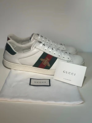 Gucci Ace vita sneakers med bi - Gucci Ace sneakers i vitt läder med klassiska gröna och röda sidoremsor samt broderad guldfärgad bi på sidan. Skorna har vita snören, grön och röd detalj på hälen och kommer med dustbag och etikett. Perfekta för dig som gillar lyxig streetstyle.