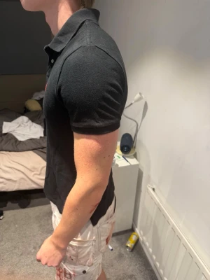 Svart pikétröja från Ralph Lauren - Klassisk svart pikétröja från Ralph Lauren med korta ärmar och röd broderad logga på bröstet. Tröjan har krage och knappar framtill. Perfekt till jeans eller shorts för en clean och stilren look.