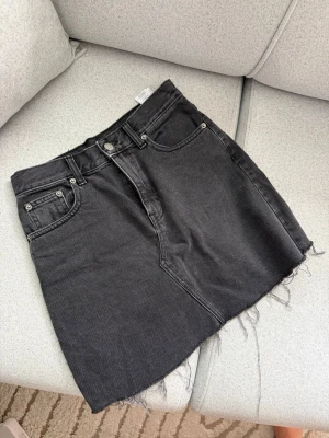 Svart jeanskjol  - Säljer en svart jeanskjol från Dropdenim med rå kant nertill och klassisk femficksdesign. Kjolen har bälteshällor och knappgylf, perfekt för en avslappnad och trendig look. Materialet är kraftig bomull med en snygg tvättad finish.
