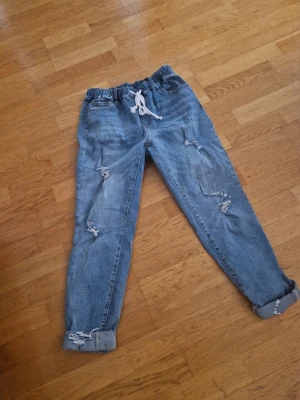 Blåa jogger jeans med dragsko - Säljer ett par ljusblå jogger jeans från shein med resår i midjan och vit dragsko. Jeansen har slitna detaljer på benen och uppvikta benslut för en avslappnad look. Perfekta för en chill och trendig stil.