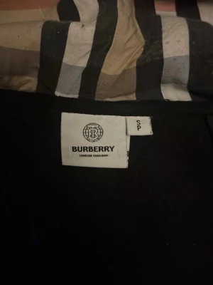 Svart hoodie från Burberry med dragkedja - Säljer en svart hoodie från Burberry med dragkedja framtill och klassiskt rutigt foder i huvan. Hoodien har långa ärmar, justerbar dragsko i huvan och ribbade muddar. Perfekt för dig som gillar stilrena och exklusiva plagg.