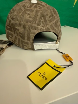 Beige keps från Fendi med FF-mönster - Snygg beige keps från Fendi med det klassiska FF-logomönstret över hela kepsen. Justerbar rem baktill i vitt läder med Fendi-logga. Kepsen är i textilmaterial och har en modern, sportig look. Perfekt för dig som gillar exklusiva accessoarer.
