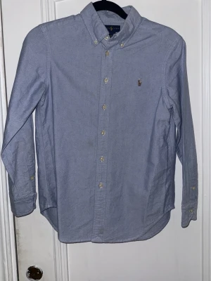 Blå skjorta från Ralph Lauren - Klassisk blå skjorta från Ralph Lauren med button-down krage och broderad logga på bröstet. Skjortan har långa ärmar, knappar framtill och är tillverkad i 100% bomull. Perfekt för en clean och stilren look.