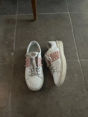 valentino skor - Säljer ett par vita sneakers från Valentino med rosa band och coola nitar på sidorna. Skorna har en chunky sula och är gjorda i läder. Perfekta för dig som vill ha en unik och trendig look med lite edge.
