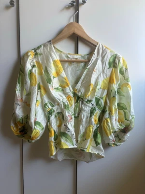 Citronmönstrad blus med puffärm - Populär blus med citronmönster i gult och grönt på vit botten. Blusen har djup v-ringning, puffiga ärmar och smock i midjan för en feminin siluett. Tillverkad i ett lätt och luftigt tyg, perfekt för sommardagar. Använd flåtal gånger och inga tecken på slitage. 