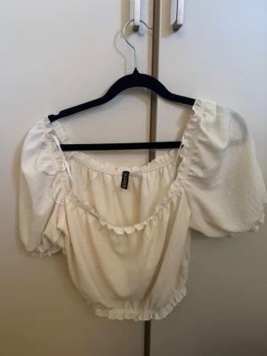 Vit puffärm blus från Chiquelle - Supersöt vit blus från Chiquelle med puffärmar och volangdetaljer vid halsringning och nederkant. Blusen har en croppad passform och är tillverkad i ett lätt, strukturerat tyg som känns luftigt och fräscht. Perfekt för dig som gillar romantisk och trendig stil.