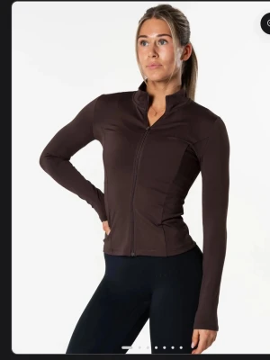 Brun träningsjacka med dragkedja - Slimmad brun träningsjacka med hel dragkedja framtill och hög krage. Tillverkad i stretchigt material som passar perfekt för träning eller uppvärmning. Långärmad modell med figurnära passform och stilren design.