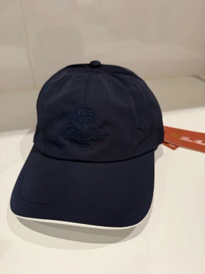 Mörkblå keps från Loro Piana - Snygg mörkblå keps från Loro Piana med broderad logga framtill. Kepsen har klassisk böjd skärm med vit kant och insida i rutigt tyg samt orange detaljer. Tillverkad i Italien med Storm System-teknologi för extra skydd mot väder.